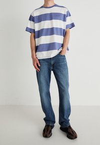 Randig t-shirt i lila och vit bomull, rund halsringning, avslappnad passform, ihop med blå denimjeans och bruna/svarta sneakers.