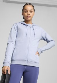 Hellblaues Zip-Hoodie mit einer Kordelzugkapuze, gerippten Bündchen und gesticktem Logo. Kombiniert mit lila Leggings und hält einen schwarzen Gegenstand.