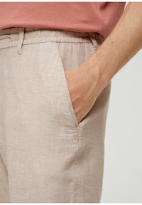 Hand resting in the side pocket of beige linen pants worn with a muted coral shirt, cropped to show lower torso and arm.