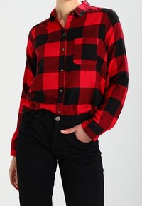 Persona vestita con una camicia a maniche lunghe a scacchi rossi e neri, con una tasca frontale, infilata in pantaloni neri a vita alta, con una mano in tasca.