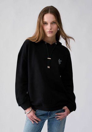 Jeune femme aux cheveux châtain clair porte un sweat à capuche noir et un jean bleu, posant avec les mains près des hanches sur un fond gris uni.
