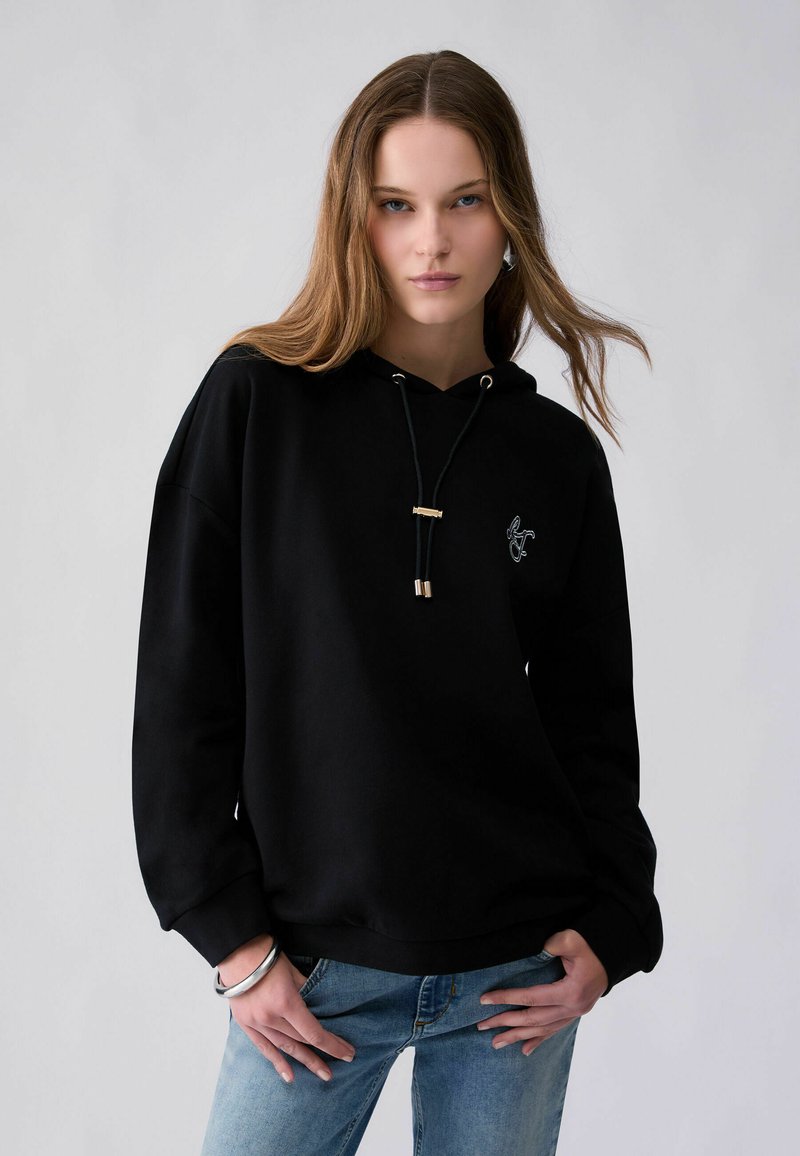 Jeune femme aux cheveux châtain clair porte un sweat à capuche noir et un jean bleu, posant avec les mains près des hanches sur un fond gris uni.