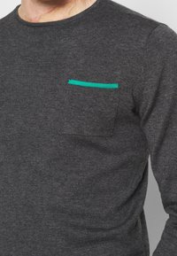 Dunkelgraues Langarmshirt aus Strickstoff mit einer Brusttasche, die mit einem kontrastierenden grünen Besatz versehen ist. Sitzt figurbetont.