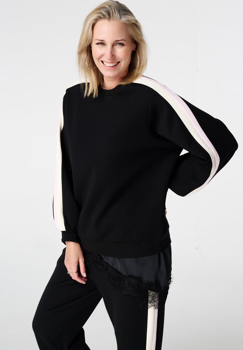 Zwarte sweatshirt met roze en witte gestreepte accenten op de mouwen, voorzien van een relaxte pasvorm en geribbelde manchetten. Gecombineerd met zwarte broek met kant.