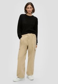 Schwarzer, gestrickter Pullover, beige Cargo-Hosen mit Seitentaschen, leichtes Material, lockere Passform, weiße Sneakers, einfarbiger Hintergrund.