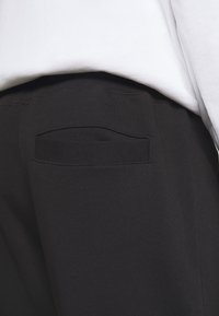 Schwarze Jogginghose aus strukturiertem Stoff, mit einer genähten Gesäßtasche und einem hohen Bund, teilweise bedeckt von einem locker sitzenden weißen Oberteil.