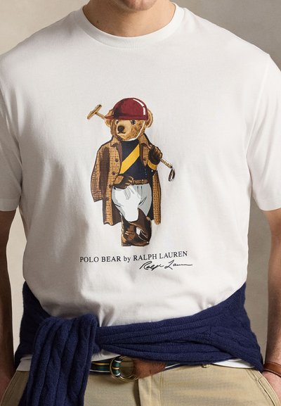 Mann trägt ein weißes T-Shirt mit einer Grafik des Polo Bears in Reitausrüstung, der ein Polomallet hält, und einem dunkelblauen Pullover, der um die Taille gebunden ist.