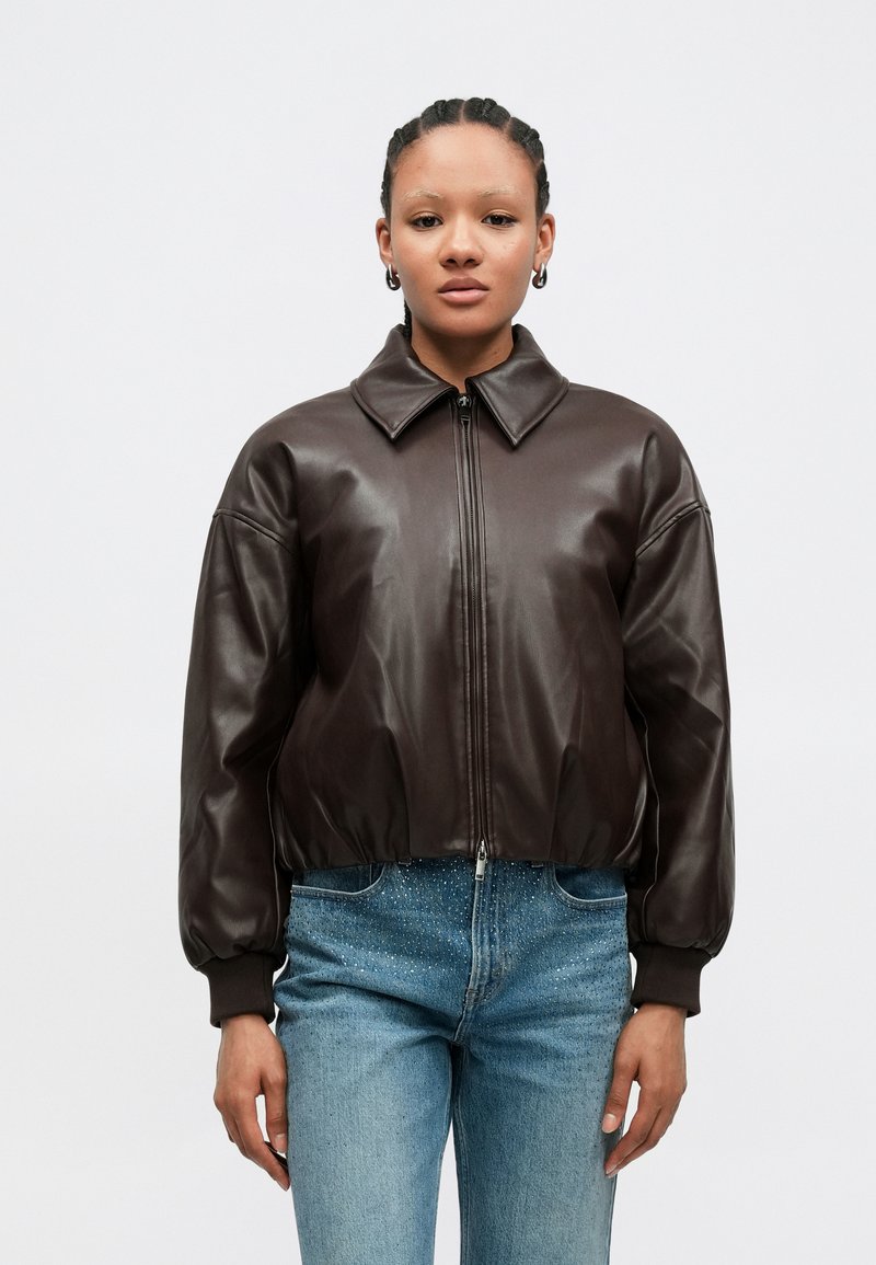 Καφέ δερμάτινο μπουφάν βόμβερ με μπροστινό φερμουάρ, μεγάλο γιακά και ελαστικά μανίκια, συνδυασμένο με ανοιχτόχρωμα τζιν παντελόνια με διακριτική λάμψη.