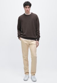 Brunstickad tröja, crèmefärgade byxor, vita sneakers. Modellen står med ena handen i fickan, enfärgad bakgrund.