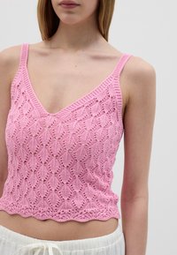 Top corto lavorato a maglia di colore rosa con scollo a V, caratterizzato da un orlo ondulato e una texture a pizzo. Spalline sottili completano il design.