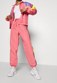 Růžová tepláková souprava se zipovou bundou s vícebarevnými rukávy, odpovídající kalhoty s bočními pruhy a elastickými manžetami. Doplněno bílými teniskami.