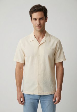 Uomo che indossa una camicia beige chiaro a maniche corte con bottoni e colletto e jeans azzurri, in piedi davanti a uno sfondo neutro.