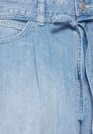 Gros plan sur un pantalon en denim bleu clair avec poche avant, passants de ceinture, une ceinture en tissu nouée et une fermeture bouton en métal.