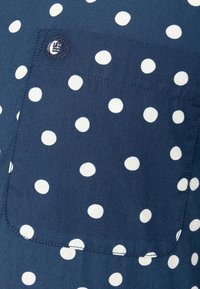 Tessuto blu navy con pois bianchi, dotato di taschino sul petto e di un bottone arrotondato. Texture liscia e dettagli di cucitura su misura.
