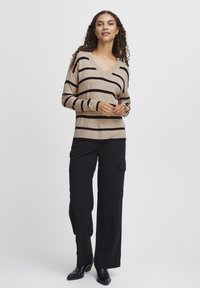 Beige gestreifter Pullover mit dunklen Streifen, V-Ausschnitt, langen Ärmeln; kombiniert mit schwarzen, weit geschnittenen Cargo-Hosen und schwarzen Stiefeletten.