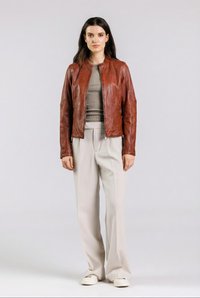 Nuori nainen seisoo ruskea nahkatakki päällä, harmaa ribbineulospaita, beiget leveälahkeiset housut ja valkoiset tennarit, yksinkertaista taustaa vasten.