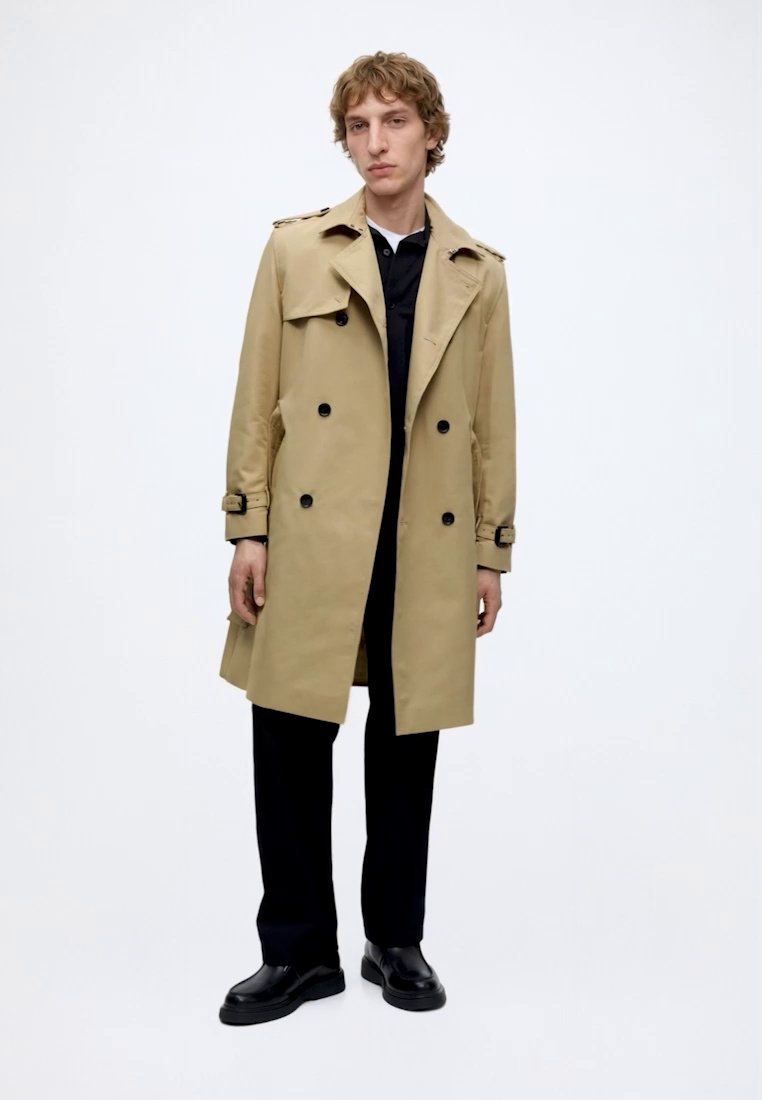 Homme portant un trench-coat beige à double boutonnage, pantalon noir, chaussures noires, debout devant un fond blanc uni.