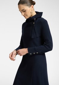 Abito blu navy con colletto alto, dotato di bottoni decorativi e un nastro in tessuto. Maniche lunghe con dettagli a bottone ai polsini.