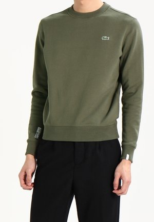 Mann trägt ein olivgrünes Sweatshirt mit Rundhalsausschnitt und langen Ärmeln mit kleinem Krokodil-Logo auf der Brust und schwarze Hose.