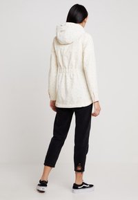 Kvinna med långt mörkt hår som bär en krämfärgad jacka med huva, svarta avklippta jeans med ett hål vid fållen, och svart-vita sneakers, står vänd bort.