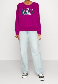 Sweat-shirt violet avec le logo "GAP" en vert foncé, associé à un jean droit bleu clair et des baskets noires. Tissu doux et décontracté.