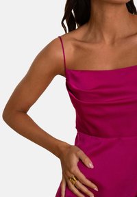 Fuchsia satijnen jurk met vierkante halslijn en dunne bandjes, met zachte draperingen en een getailleerd silhouet. Gouden ringaccent aan de hand.