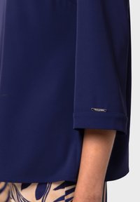 Blusa blu navy con maniche a tre quarti, realizzata in un materiale morbido. Presenta un'etichetta con accento argentato sull'orlo delle maniche.