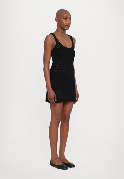 Rochie tricotată neagră cu un design ajustat, având un decolteu rotunjit și un model perforat pe întreaga suprafață. Modelul stă îmbrăcat cu balerini negri.