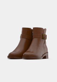 Smeđe kožne gležnjače s okruglim vrhom, bočnim patentnim zatvaračem i ukrasnim remenom sa zlatnim kopčom. Ravna potpetica. Glatka tekstura.