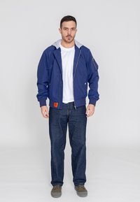 Marineblauwe bomberjack met een grijze hoodie-voering, ritssluiting aan de voorkant, elastische manchetten en zijzakken, gedragen met donkerblauwe jeans en grijze schoenen.