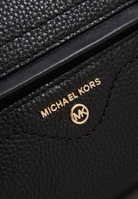 Superficie in pelle nera testurizzata con logo Michael Kors metallico dorato ed emblematico cerchio MK su un pannello cucito.