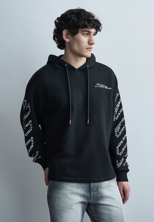 Νεαρός άνδρας με σγουρά μαλλιά που φοράει μαύρο φούτερ με λευκό κείμενο στα μανίκια και στο στήθος, ανοιχτό γκρι τζιν, κοιτάζοντας δεξιά μπροστά σε μονόχρωμο φόντο.