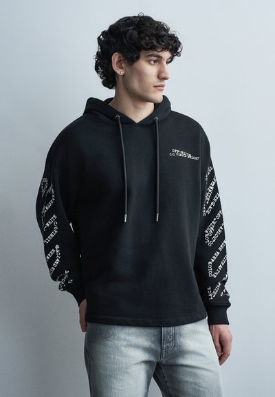 Jeune homme aux cheveux bouclés portant un sweat à capuche noir avec texte blanc sur les manches et la poitrine, jean gris clair, faisant face à droite sur un fond uni.