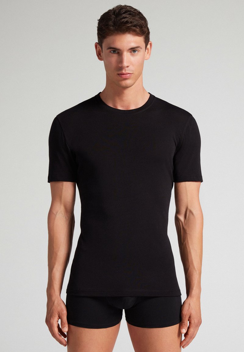 Junger Mann steht frontal, trägt ein eng anliegendes schwarzes Kurzarm-T-Shirt und schwarze Boxershorts vor einfarbigem Hintergrund.