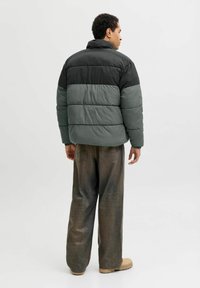 Pufferjacka i svart och grönt med horisontella segment, hög krage och elastiska muddar. Matchas med lösa, blekta bruna byxor.