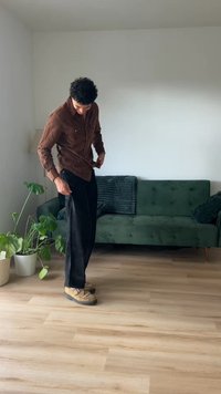 Hnědá košile na knoflíčky, černé široké kalhoty, béžové boty s dekorativním designem. Zelená pohovka a rostliny v pozadí, dřevěná podlaha.