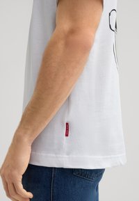 Weißes Baumwoll-T-Shirt mit kurzen Ärmeln, das mit einem grafischen Druck versehen ist. Sichtbares rotes Etikett an der Seitennaht, kombiniert mit blauen Jeans.