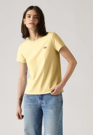 Jeune femme portant un t-shirt jaune Levi's et un jean bleu clair, debout devant un fond gris clair uni.