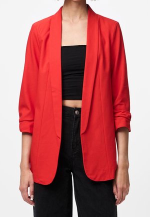 Person trägt einen leuchtend roten offenen Blazer über einem schwarzen Bandeau-Top und hoch taillierter schwarzer Hose, stehend vor einem weißen, schlichten Hintergrund.