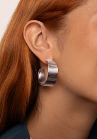 Pendiente de aro de metal plateado con un diseño curvado, que presenta una superficie brillante y una textura suave, llevado en la oreja.