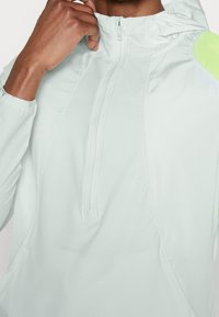 Veste coupe-vent vert menthe clair avec une demi-fermeture éclair, présentant un tissu texturé et un accent contrastant en vert lime vif sur l'épaule.