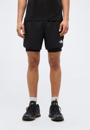 Άνδρας που φοράει μαύρα στρωμένα σορτς για τρέξιμο, μαύρα αθλητικά παπούτσια, γκρι κάλτσες και μαύρο πουκάμισο, στέκεται σε ένα απλό ανοιχτό φόντο.