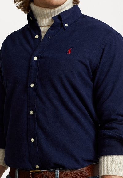 Camisa azul-marinho com botões brancos, apresentando um logótipo bordado em vermelho. Usada sobre um suéter de gola alta creme, exibindo um tecido texturizado.