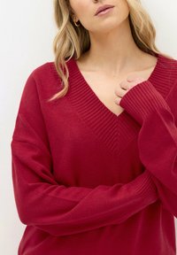 Roter Pullover mit V-Ausschnitt Design, geripptem Kragen und Bündchen. Weiche Textur, lockere Passform, mit überschnittenen Schultern und langen Ärmeln.