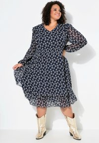 Vestido azul marino con estampado floral, mangas largas transparentes, escote en V y dobladillo con volantes, combinado con botas vaqueras metálicas doradas.