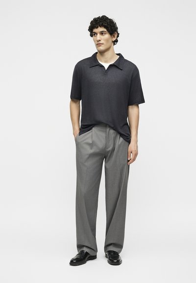 Άνδρας με κατσαρά μαλλιά, φορώντας μαύρο πόλο με κοντά μανίκια, γκρι φαρδιά παντελόνια και μαύρα παπούτσια, στέκεται με τα χέρια στις τσέπες.