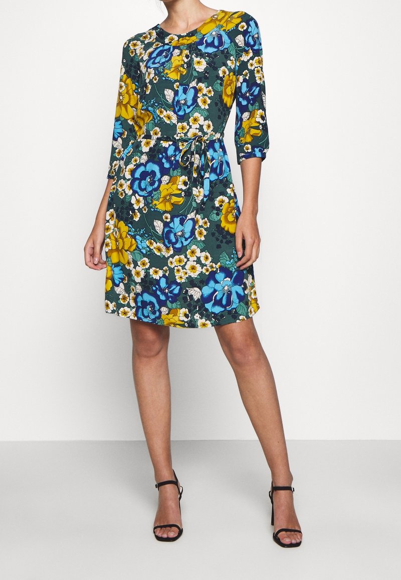 Femme portant une robe fleurie mi-longue avec des fleurs bleues, jaunes et blanches, manches trois-quarts, et des talons noirs avec bride à la cheville.