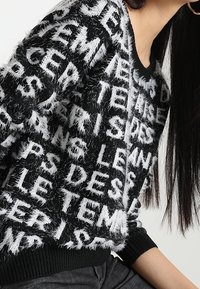 Pull en tissu flou noir et blanc avec des motifs de texte blanc audacieux. Le tissu a une finition texturée, avec un ourlet noir à côtes.