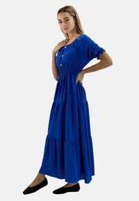 Blauwe maxi-jurk met korte pofmouwen, elastische tailleband en geplooide volantzoom. Gestyled met zwarte platte schoenen.