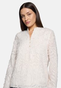 Gesprenkelte weiße Bluse mit Mandarin-Kragen, die ein dezentes Muster und eine Vorderseite mit drei Knöpfen aufweist. Lange, leicht aufgebauschte Ärmel.